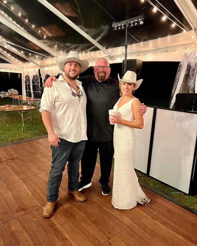 Gary Lucca with a bride and groom in cowboy hats inside a clear-top tent with bistro lights